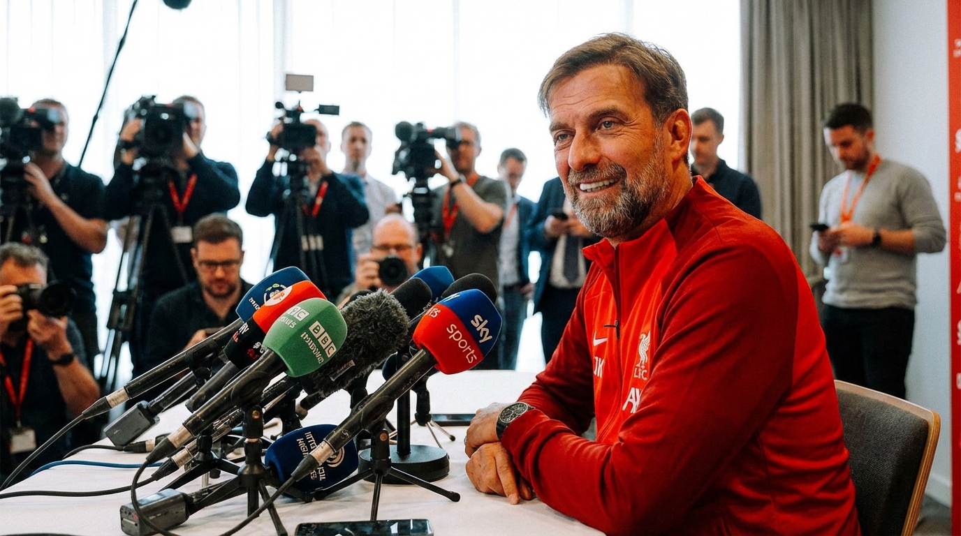 A football manager speaking into microphones at a press conference table wearing a red tracksuit jacket